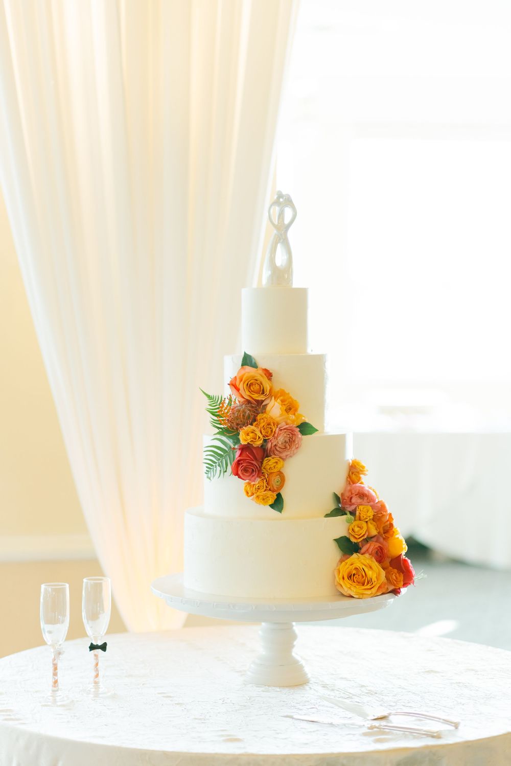 White tiered wedding cake decorated with orange roses and tropical greenery on white tablecloth.