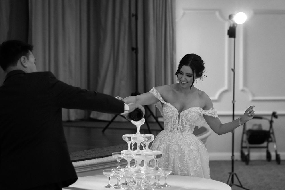 A couple pours champagne into a tower of glasses at their wedding reception in black and white.