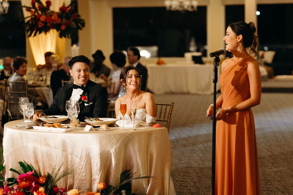 Reception scene with guests at table listening to speech in elegant ballroom setting.