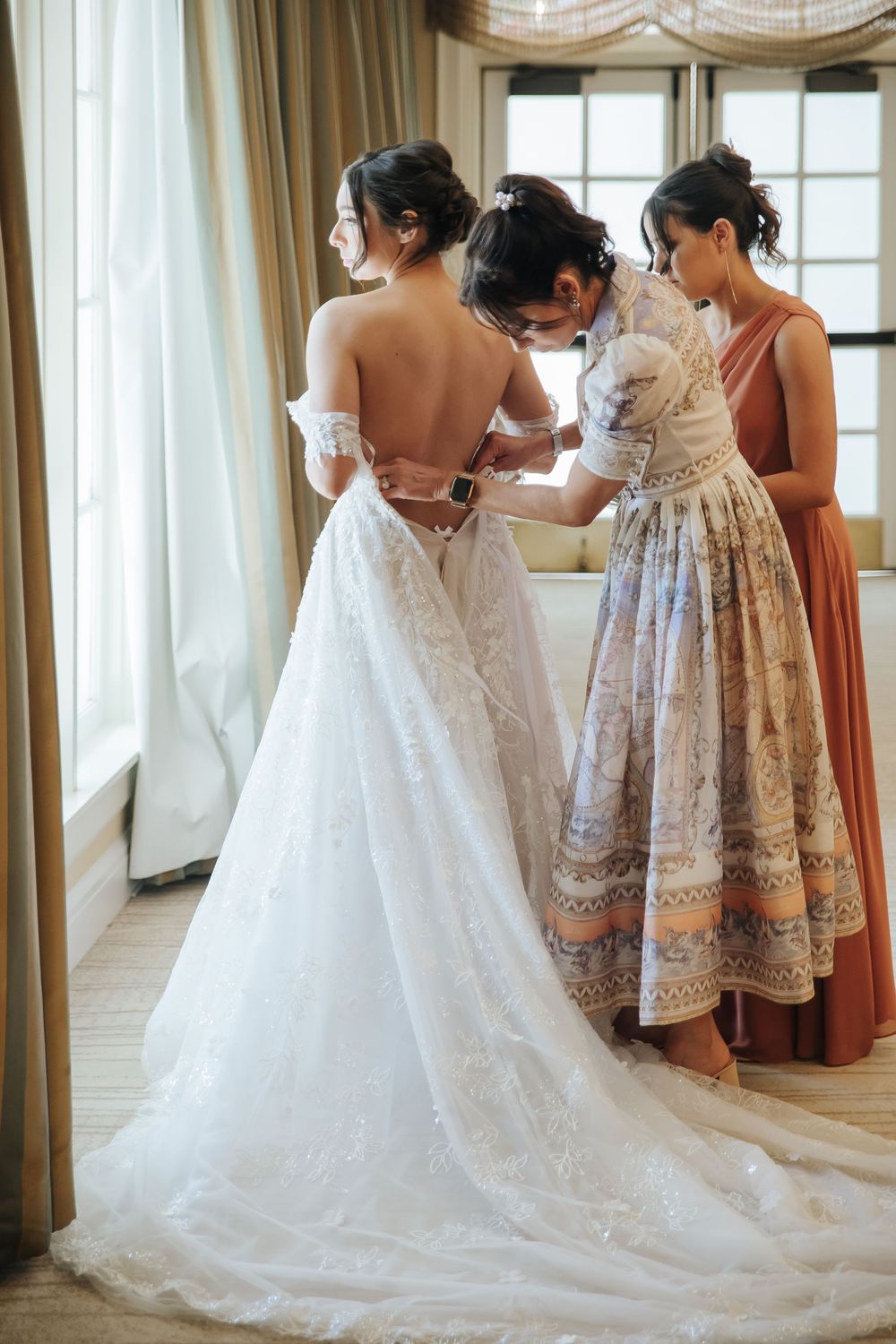 Wedding party helps with final touches on a bride's lace gown with long train near bright windows.