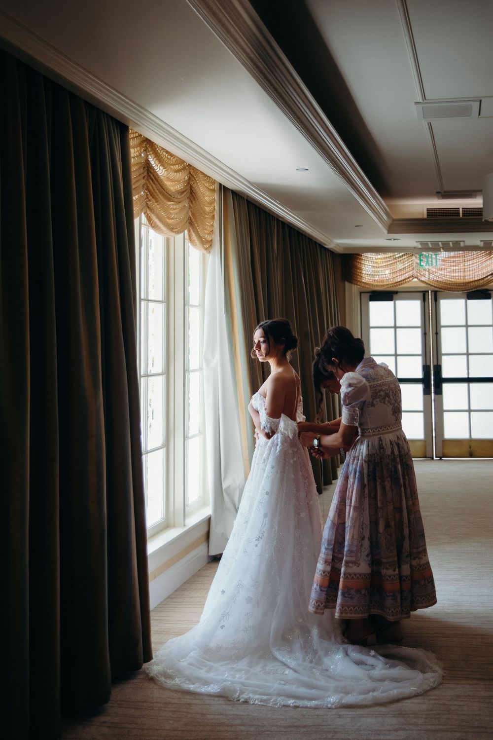 Wedding scene shows a bride in flowing white gown being helped with final preparations in an elegant room.