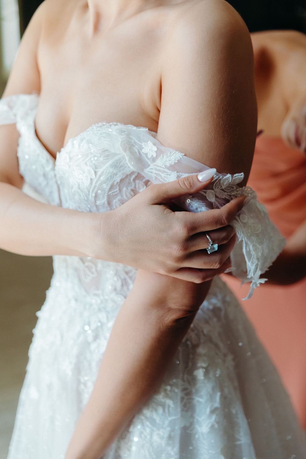 Close-up detail of white lace wedding dress with delicate embroidery and ring-adorned hand adjusting the fabric.