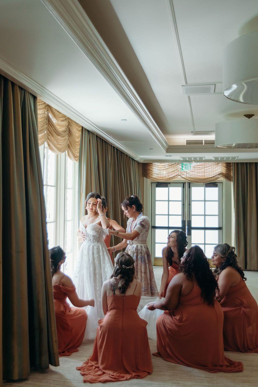 Bridesmaids in coral dresses helping with bridal preparations in a sunlit hotel room.
