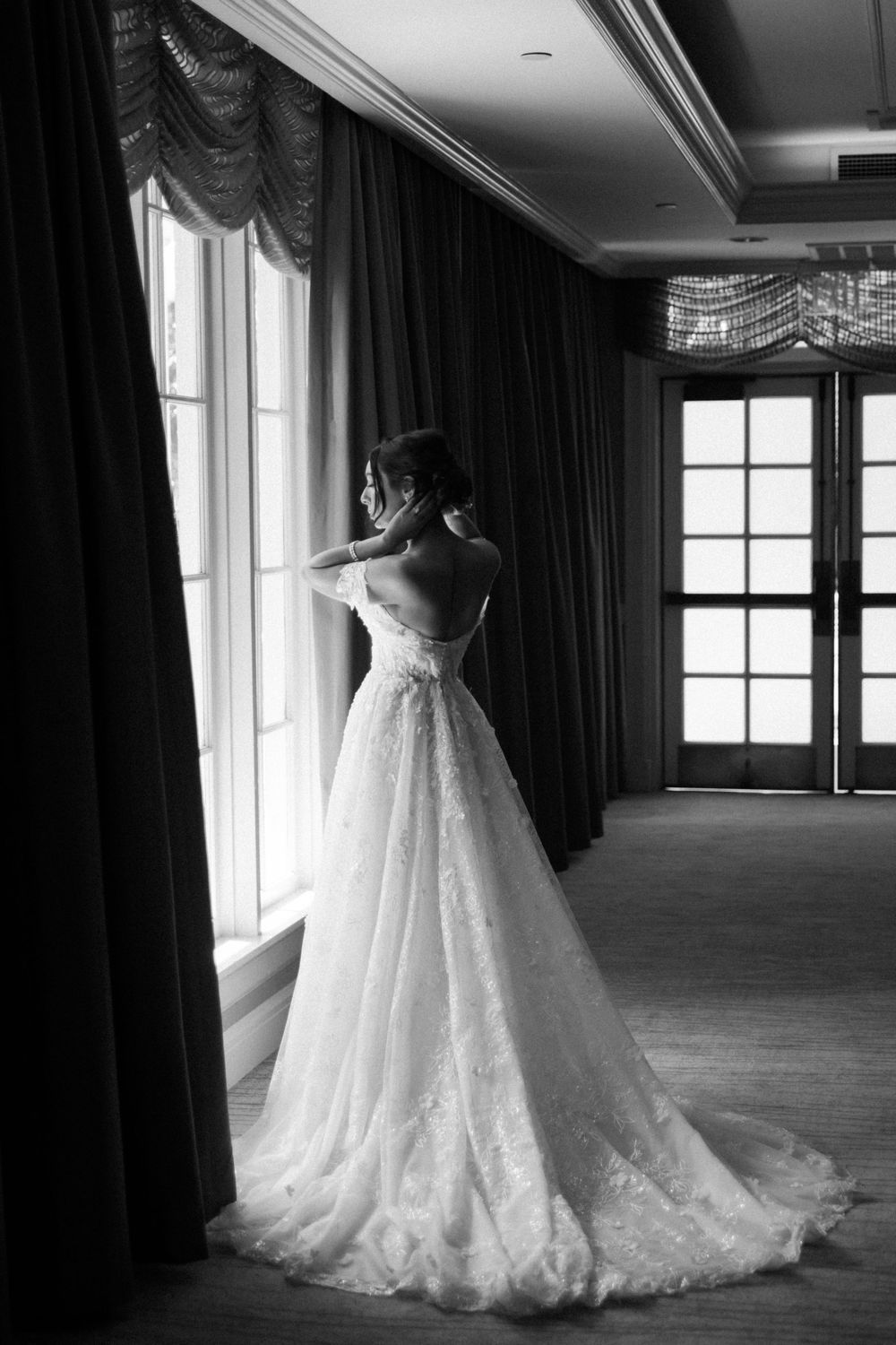 A bride in an elegant white lace wedding gown with a flowing train looks out a window in black and white.