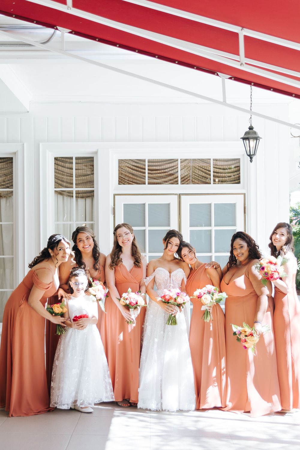 Bridesmaids wearing flowing peach-colored dresses pose together with floral bouquets outside a white building.