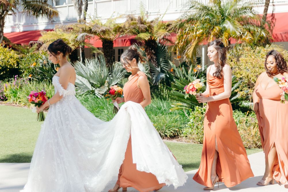 A wedding party in sunset orange dresses walks on a garden path with palm trees and tropical landscaping.