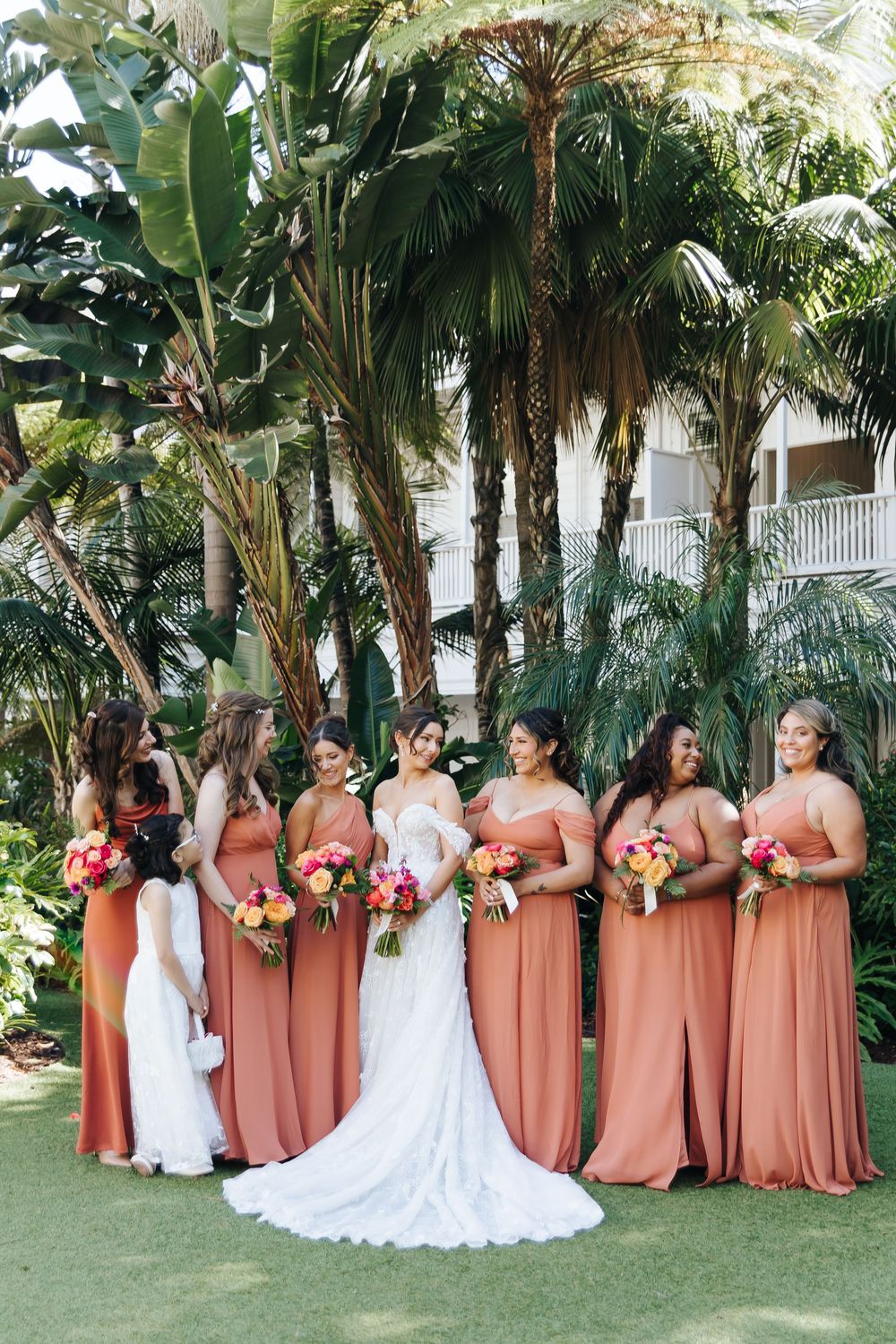 Wedding party in peachy-coral dresses pose with bouquets against a tropical palm backdrop in an outdoor garden setting.