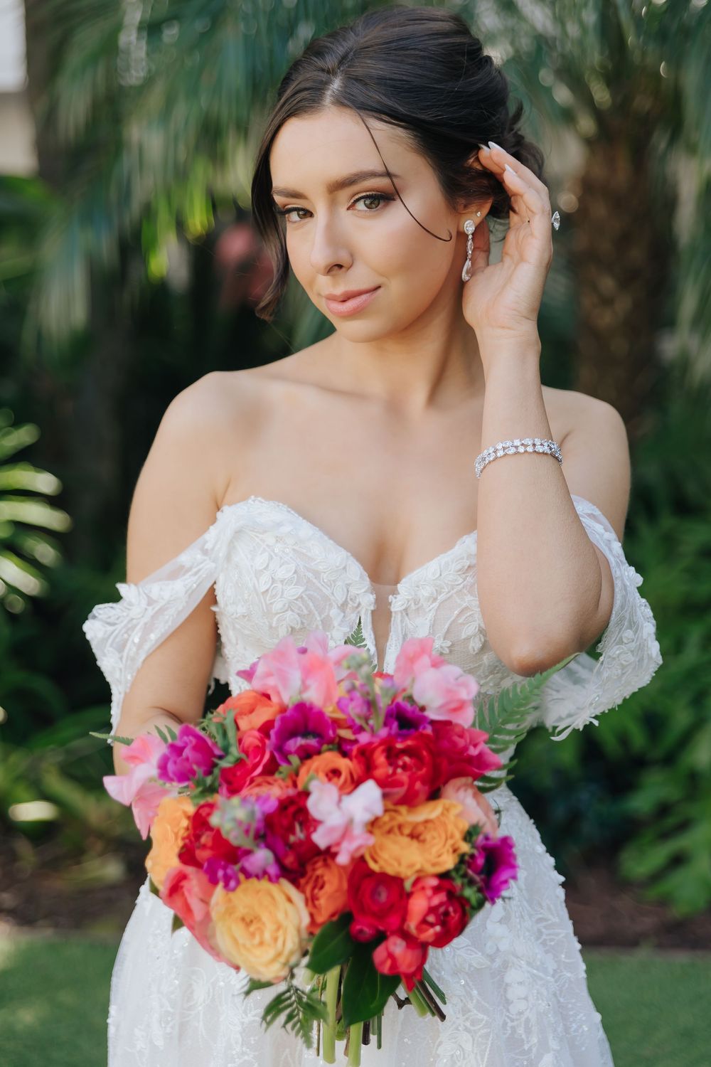 A bride in a white lace off-shoulder dress holds a vibrant bouquet of pink, orange and yellow flowers against a tropical backdrop.