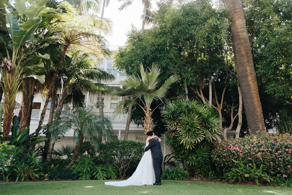 A couple embraces in front of a tropical garden with palm trees and lush greenery outside a white house.