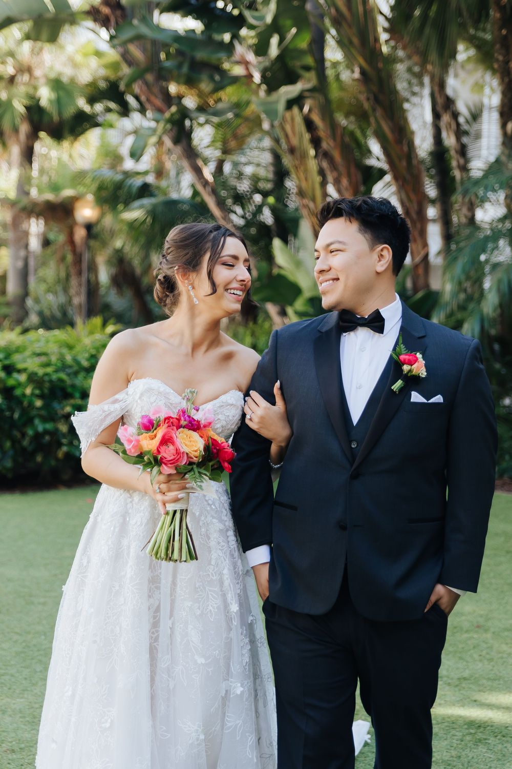 Wedding couple shares a joyful moment outdoors with pink bouquet against lush tropical greenery.