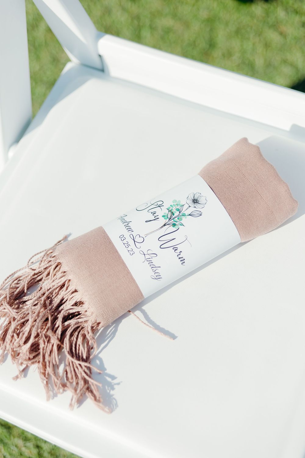 A blush pink pashmina scarf with fringe tassels wrapped in a wedding favor label rests on a white chair.