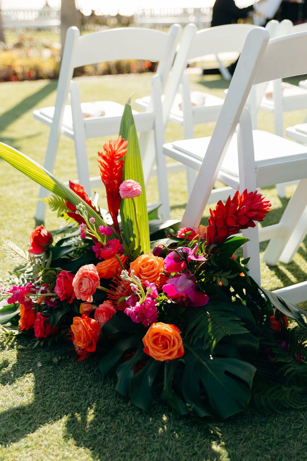 Vibrant tropical floral arrangement with red ginger and orange roses decorating white ceremony chairs.