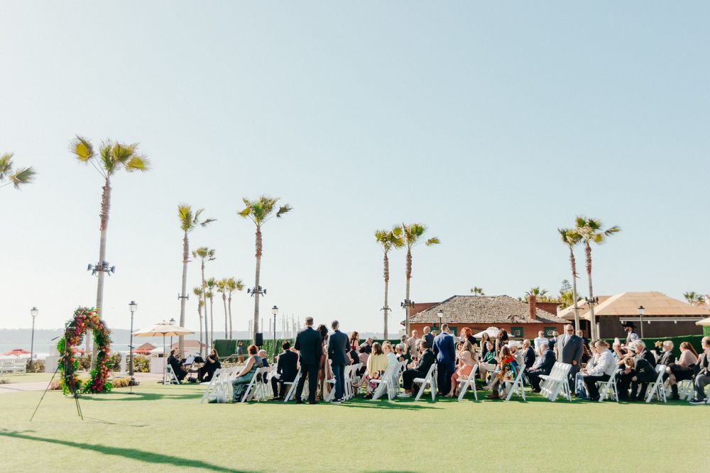 Wedding ceremony guests gather outdoors at a beachside venue with palm trees and ocean views on a sunny day.