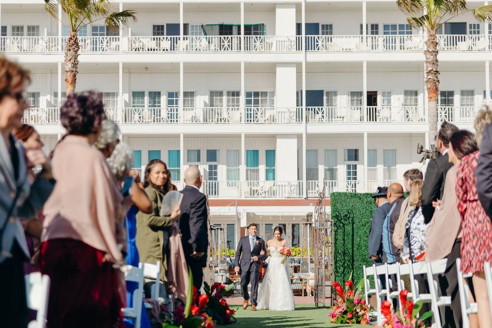 Outdoor wedding ceremony taking place on a lawn in front of a white beachfront hotel with palm trees.