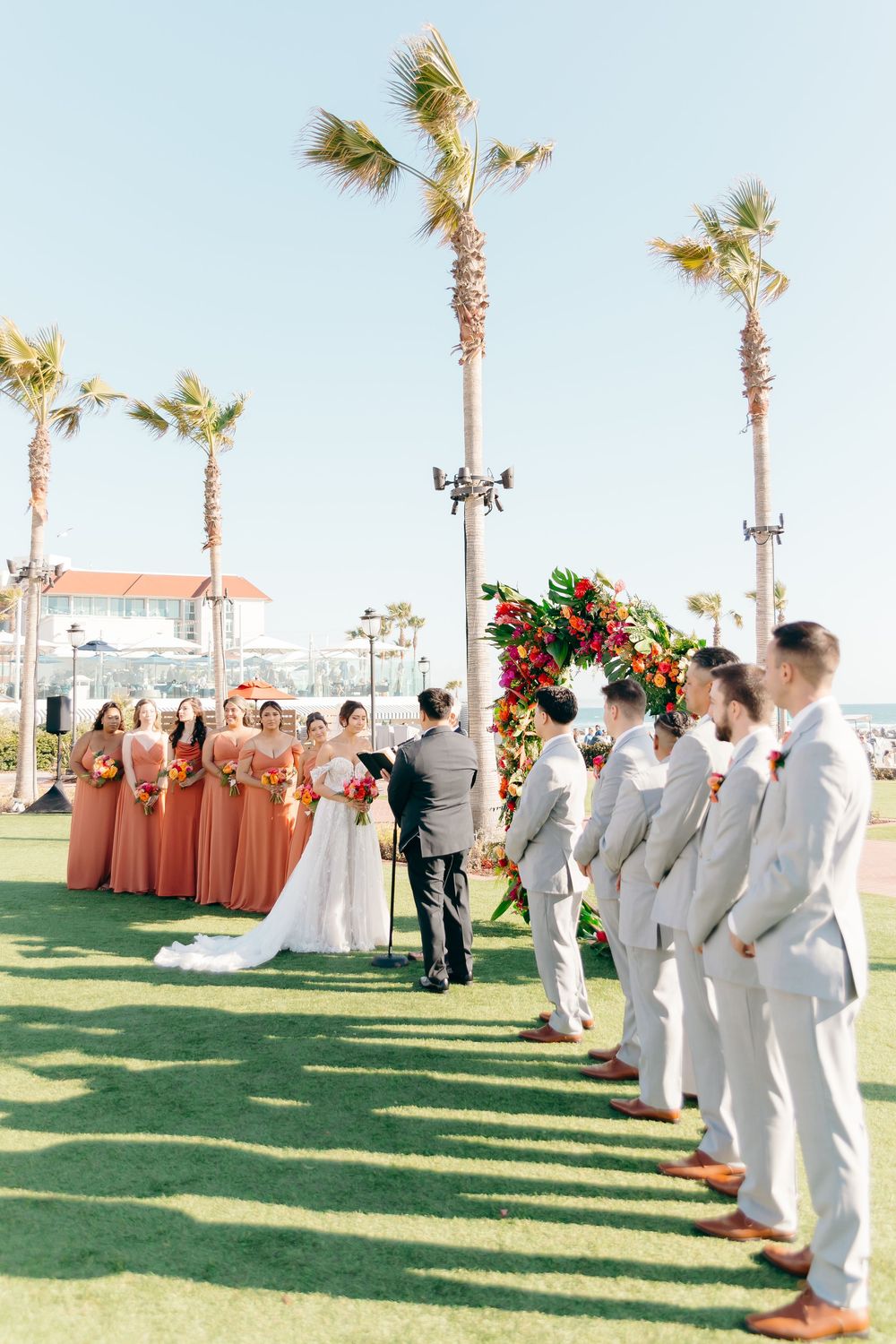 Outdoor wedding ceremony setup with palm trees, featuring bridesmaids in coral dresses and groomsmen in white suits.