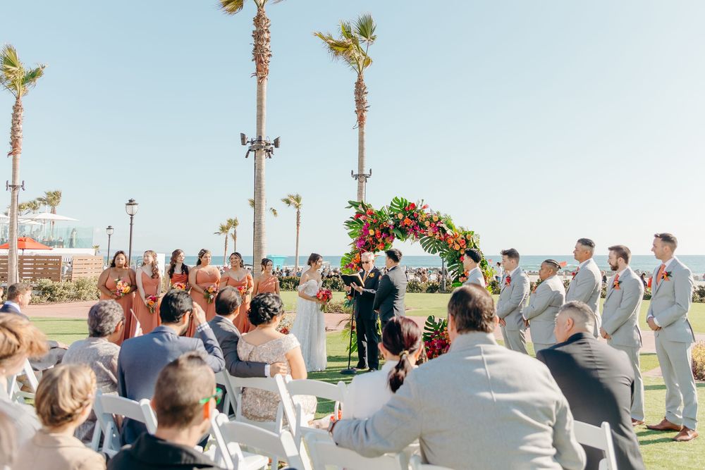 Outdoor beachside wedding ceremony with palm trees and guests seated in white chairs on a sunny day.