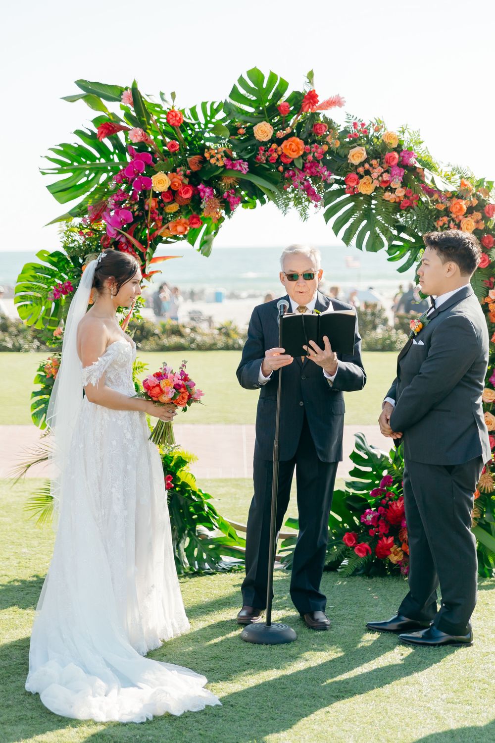 A wedding ceremony under a tropical floral arch with monstera leaves and colorful flowers on a grassy outdoor venue.