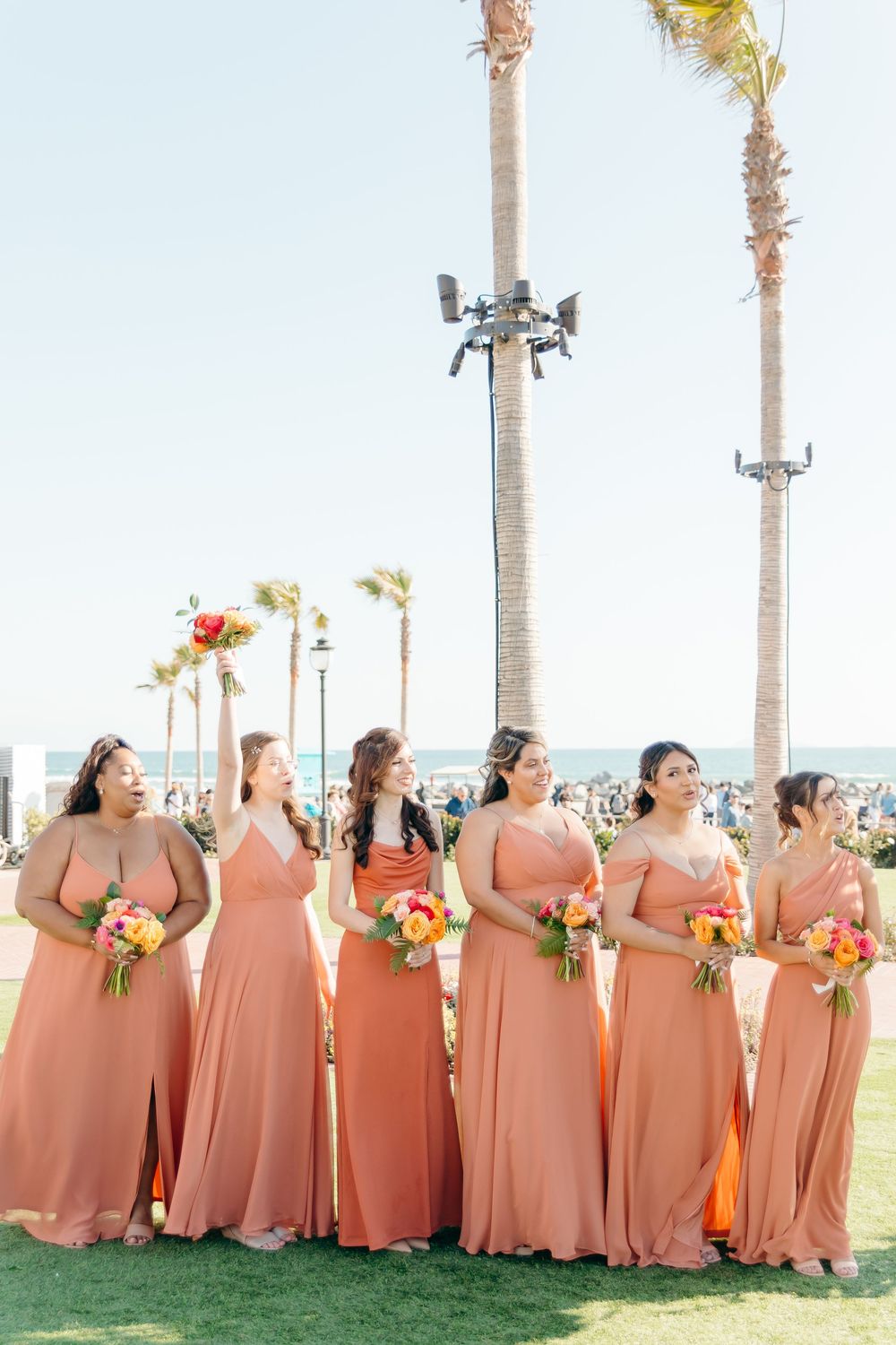 Bridesmaids in matching coral-colored dresses stand in a row outdoors with palm trees and floral bouquets.