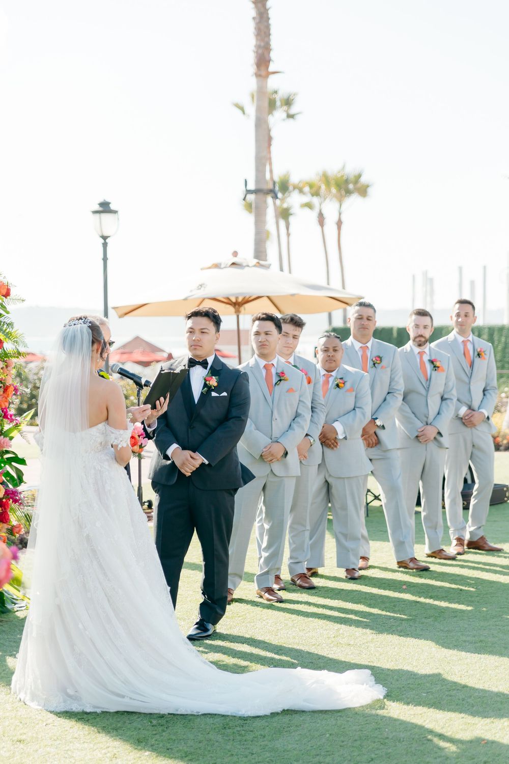 Wedding ceremony on a lawn with groomsmen in light gray suits and parasol backdrop.