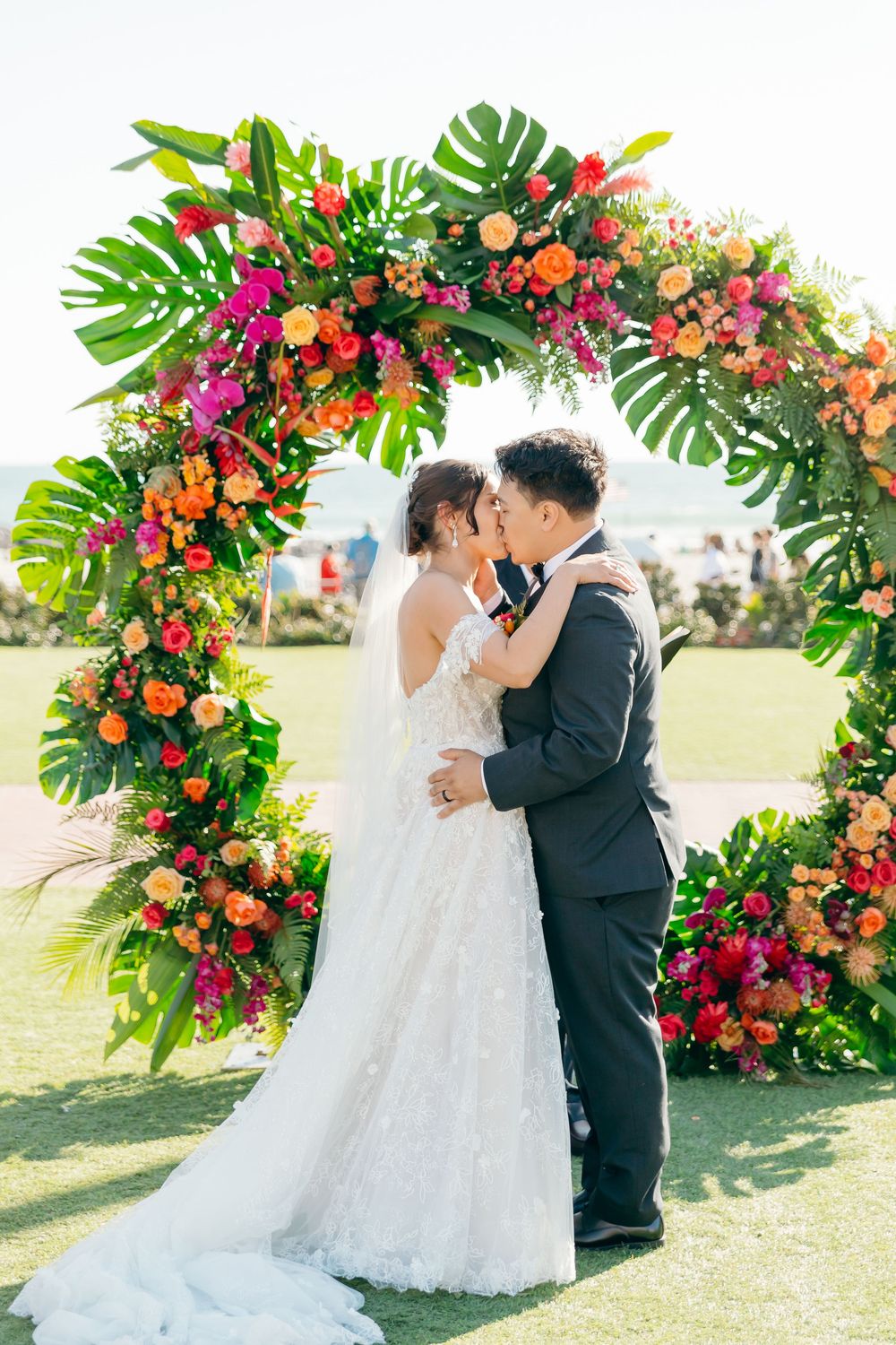 Wedding ceremony under a vibrant tropical floral arch with pink and orange blooms against a sunny backdrop.