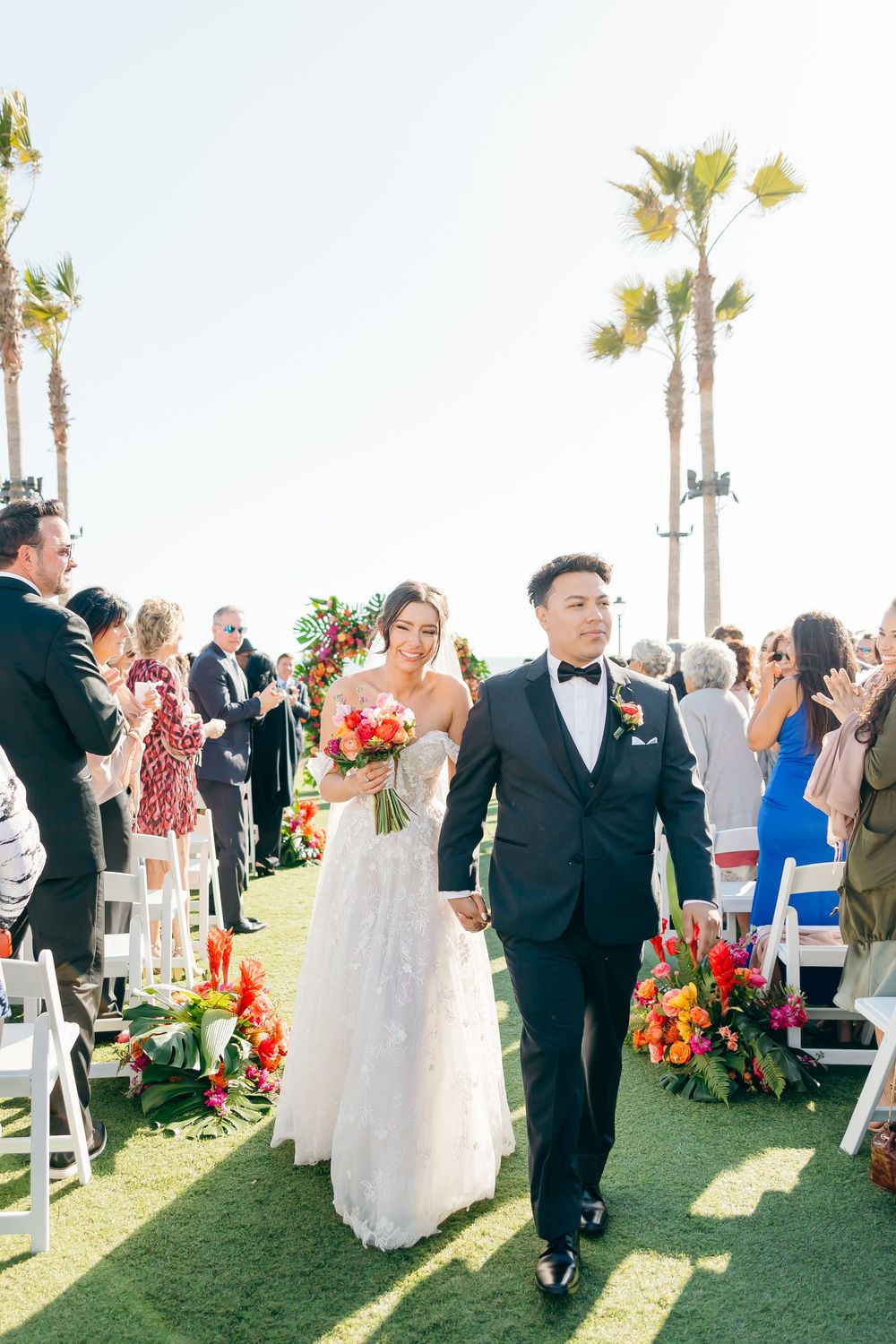 Newlyweds walking down the aisle surrounded by guests and tropical floral arrangements.