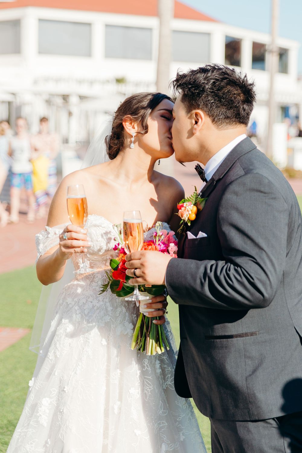 Newlyweds share a romantic kiss while holding champagne glasses and a bouquet during their outdoor wedding celebration.