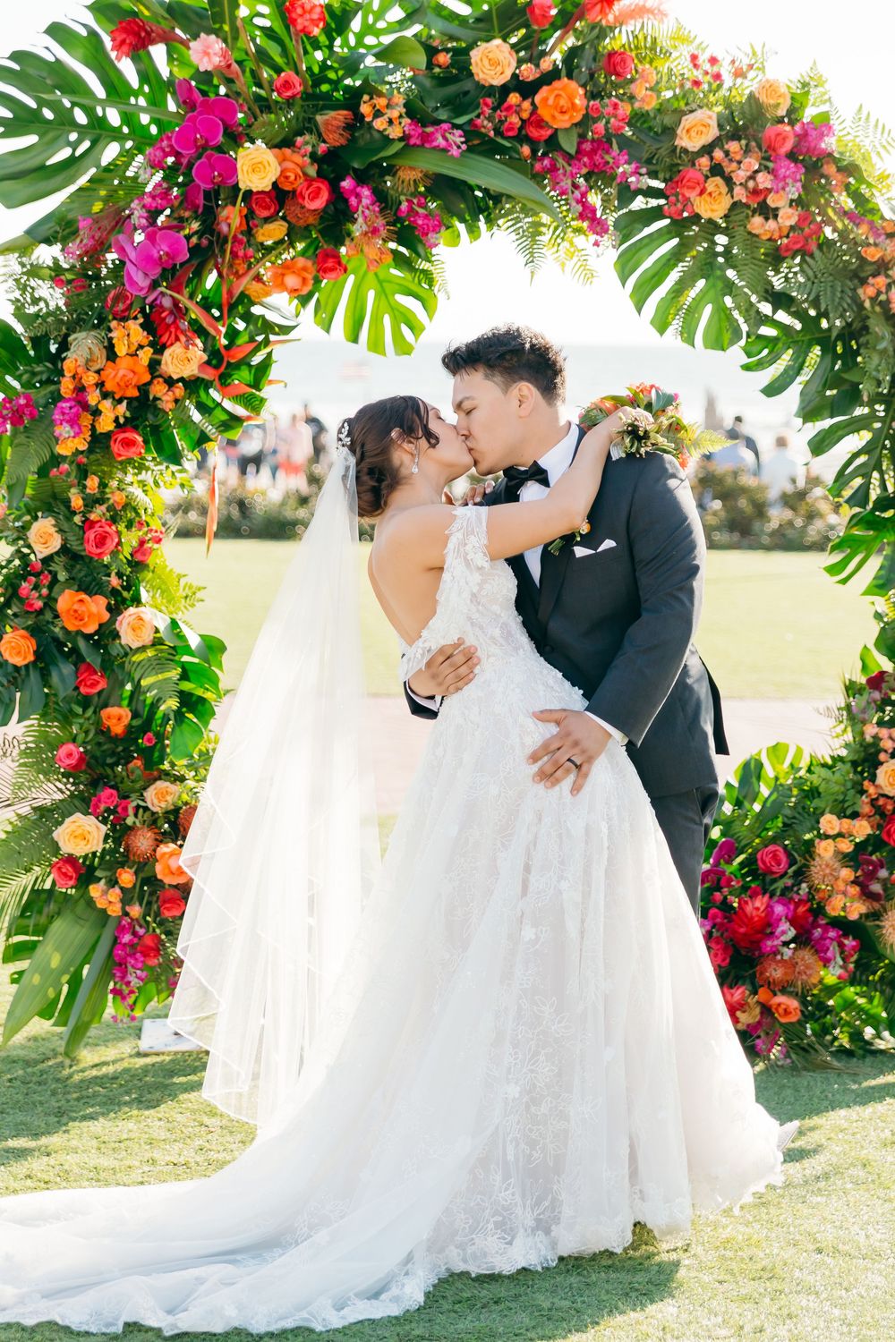 Tropical floral arch with bright flowers and monstera leaves frames a wedding couple.