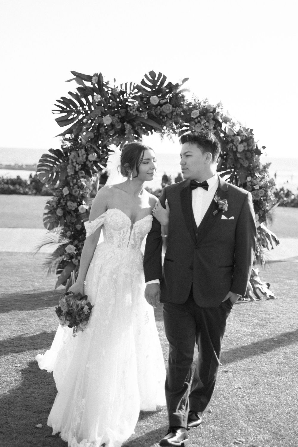 A black and white portrait of newlyweds under a tropical floral arch.