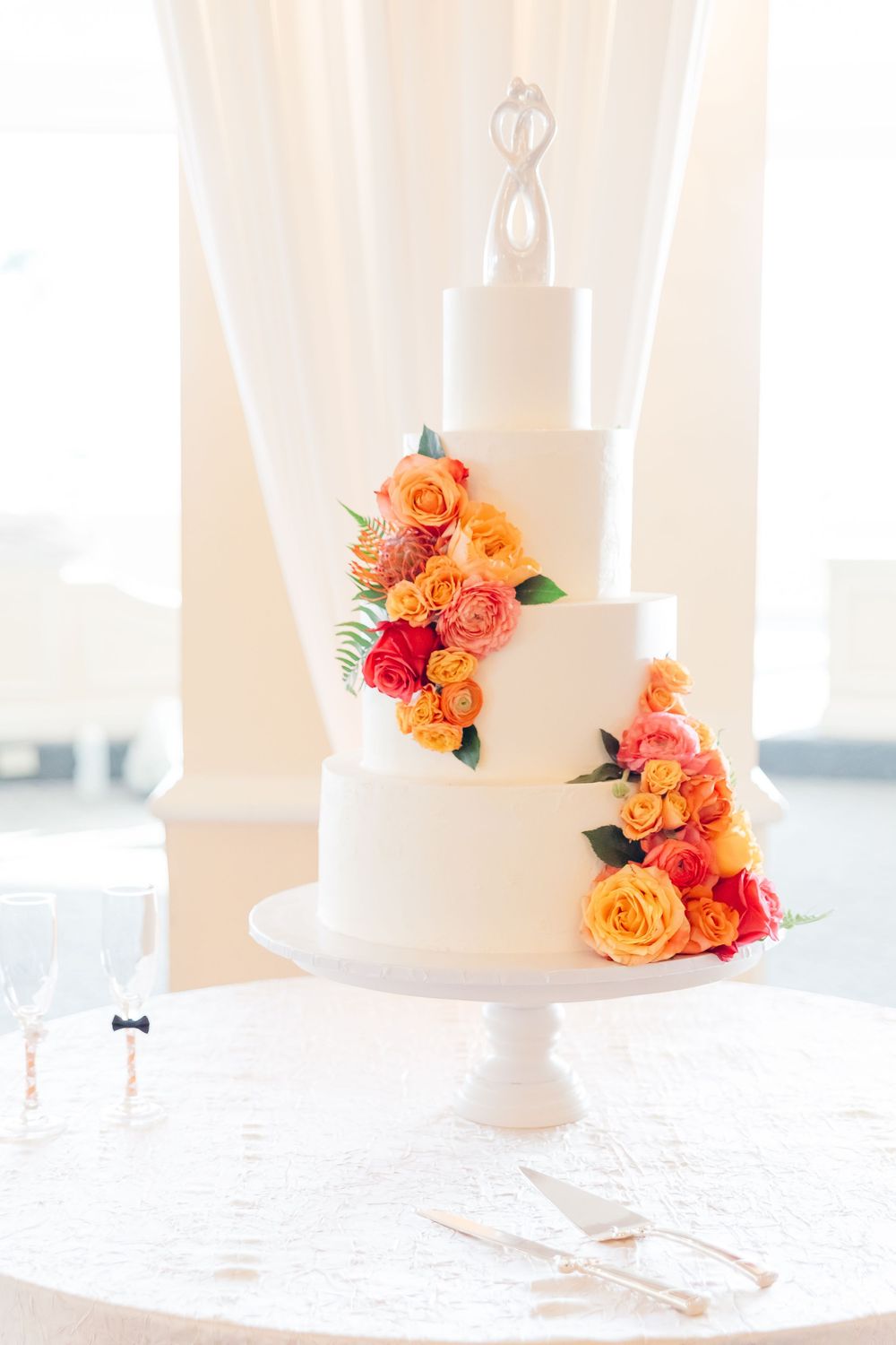 Elegant white tiered wedding cake decorated with vibrant coral and orange roses on a white stand.