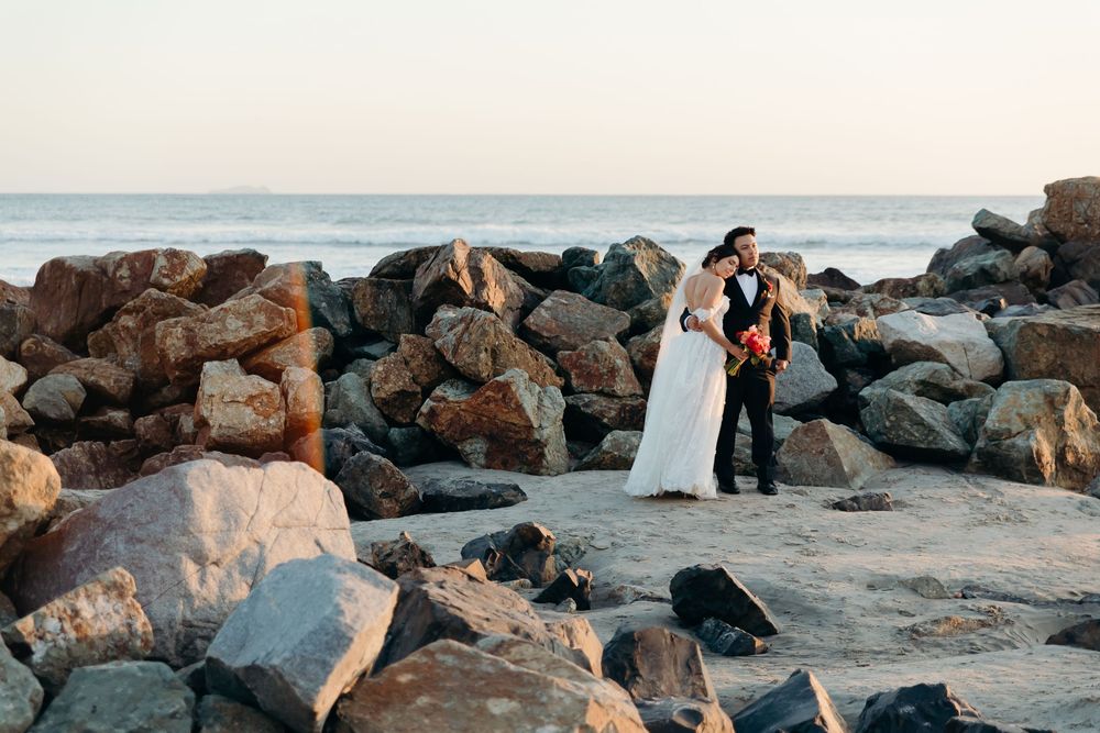 Newlywed couple embraces on rocky beach at sunset with ocean waves in background.
