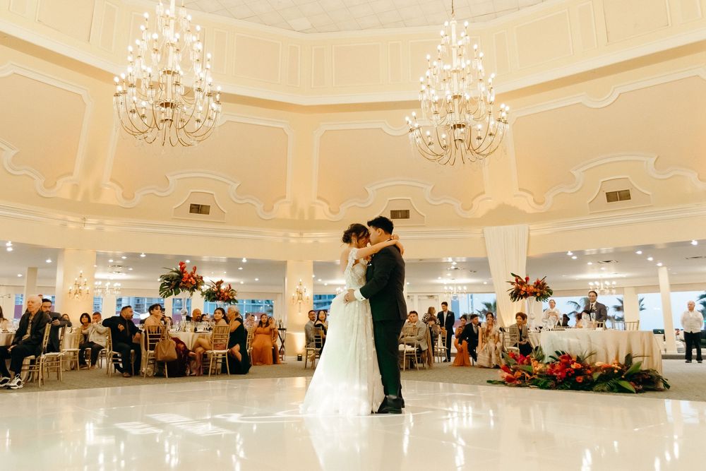 A couple shares their first dance in an elegant ballroom with crystal chandeliers and wedding guests seated at round tables.
