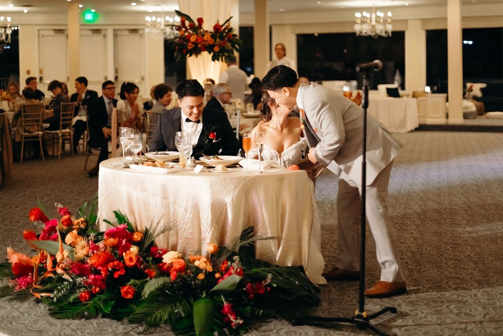 Newlyweds seated at their reception table with bright red floral arrangements.
