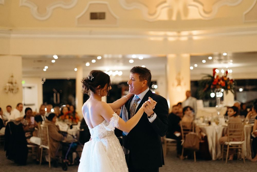 A couple shares their first dance in an elegant ballroom with ornate ceiling details and soft lighting.
