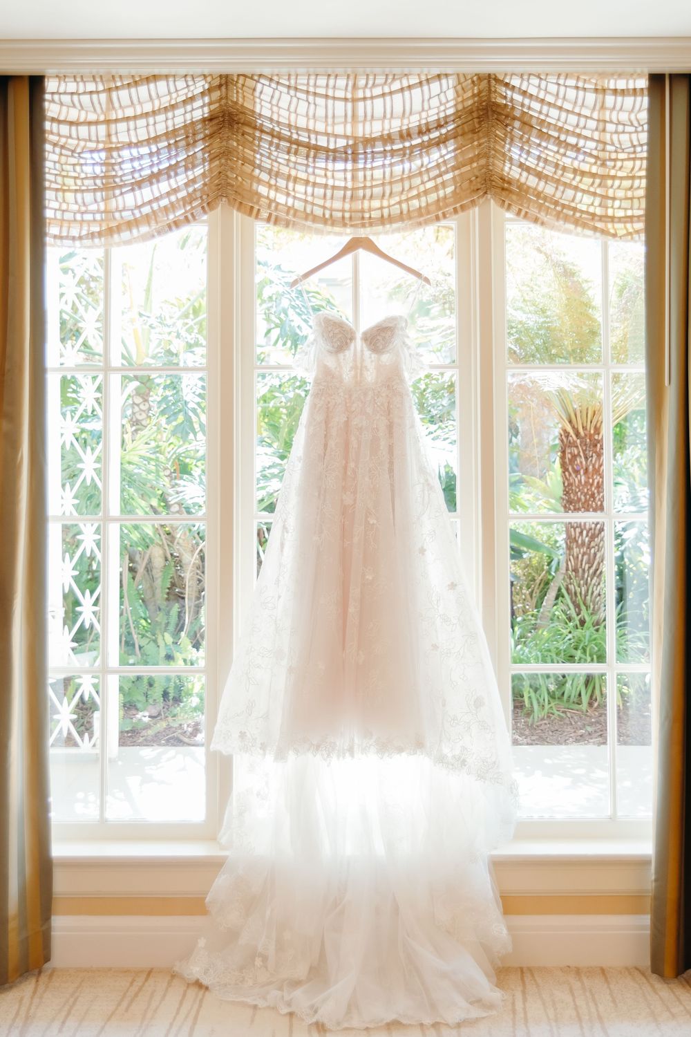 A white wedding dress hangs elegantly in front of a window with sheer curtains and tropical garden views.