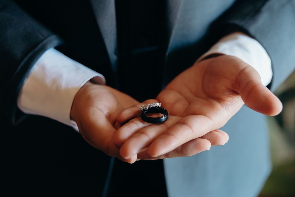 A person in a suit jacket holds a black wedding ring in their open palm against a dark background.