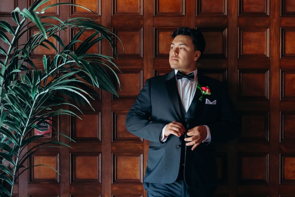 A person in a tuxedo stands against a dark wood paneled wall with a decorative plant in elegant formal attire.