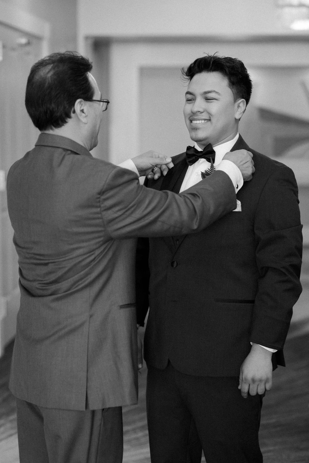 Black and white photo of a man in a suit helping to adjust another man's bow tie and formal attire.