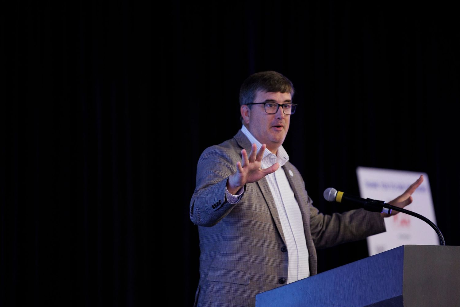 Speaker in gray blazer gestures while presenting at a podium during a professional conference.