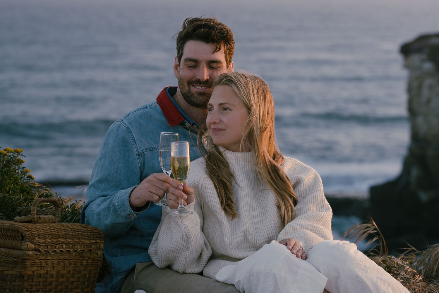 A couple shares a romantic moment with champagne on a coastal cliff at sunset while wearing cozy sweaters.