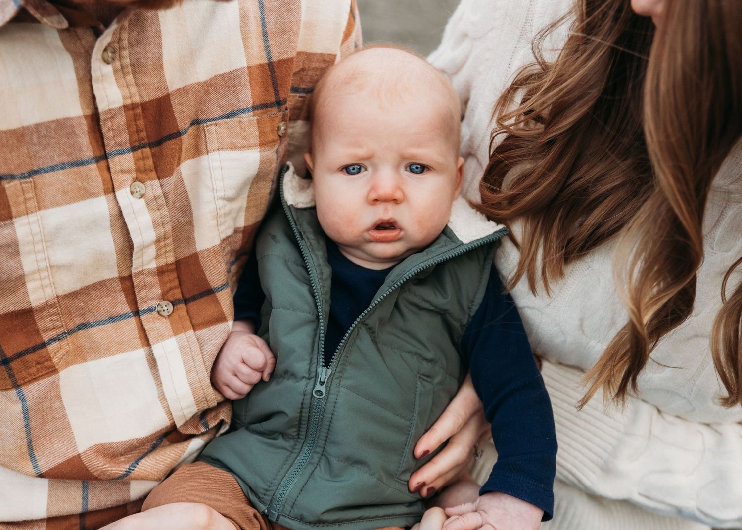A small baby wearing a green vest sits between two adults in cozy sweaters.