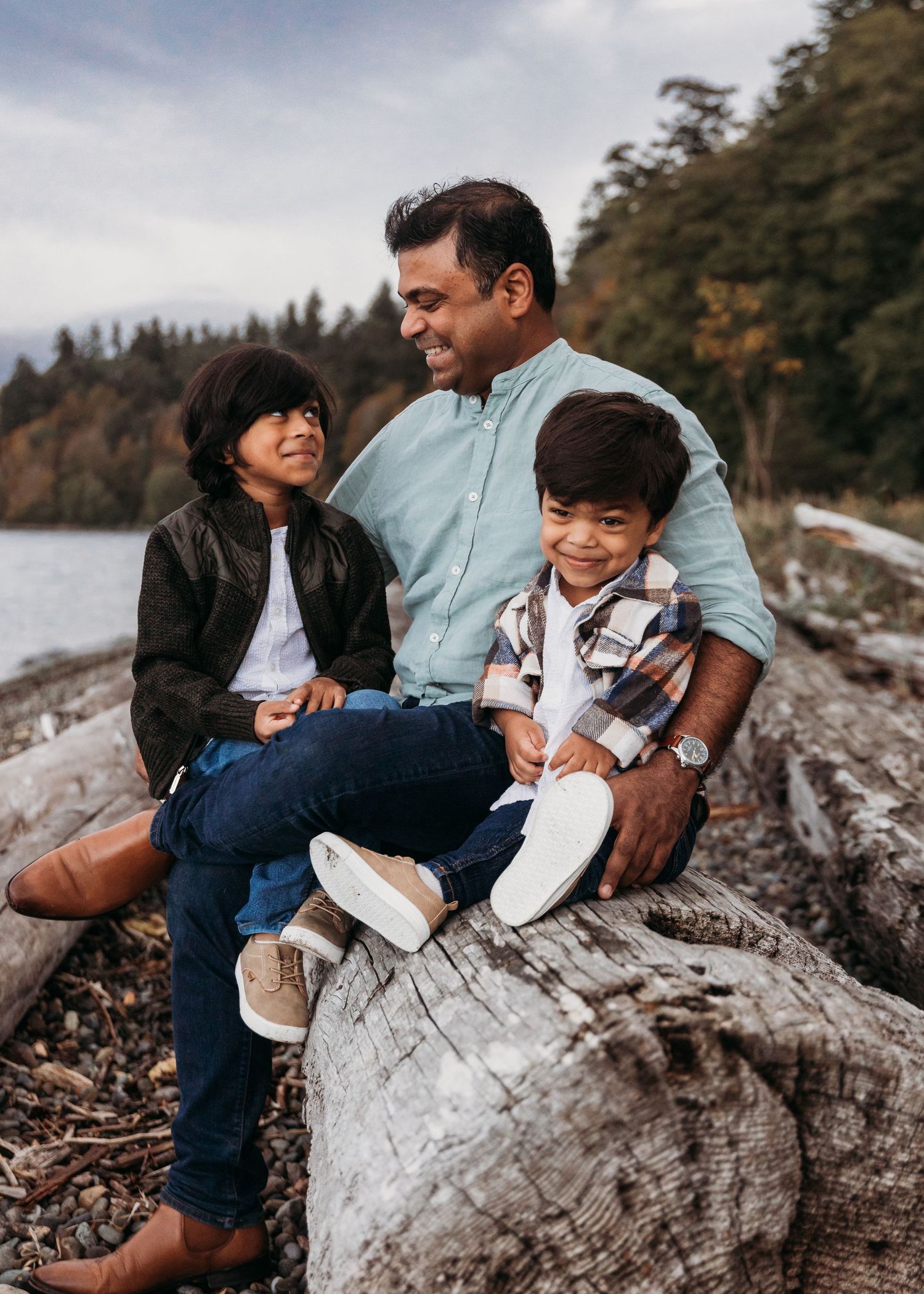 A family sits together on a large driftwood log at a rocky beach with evergreen trees in the background.