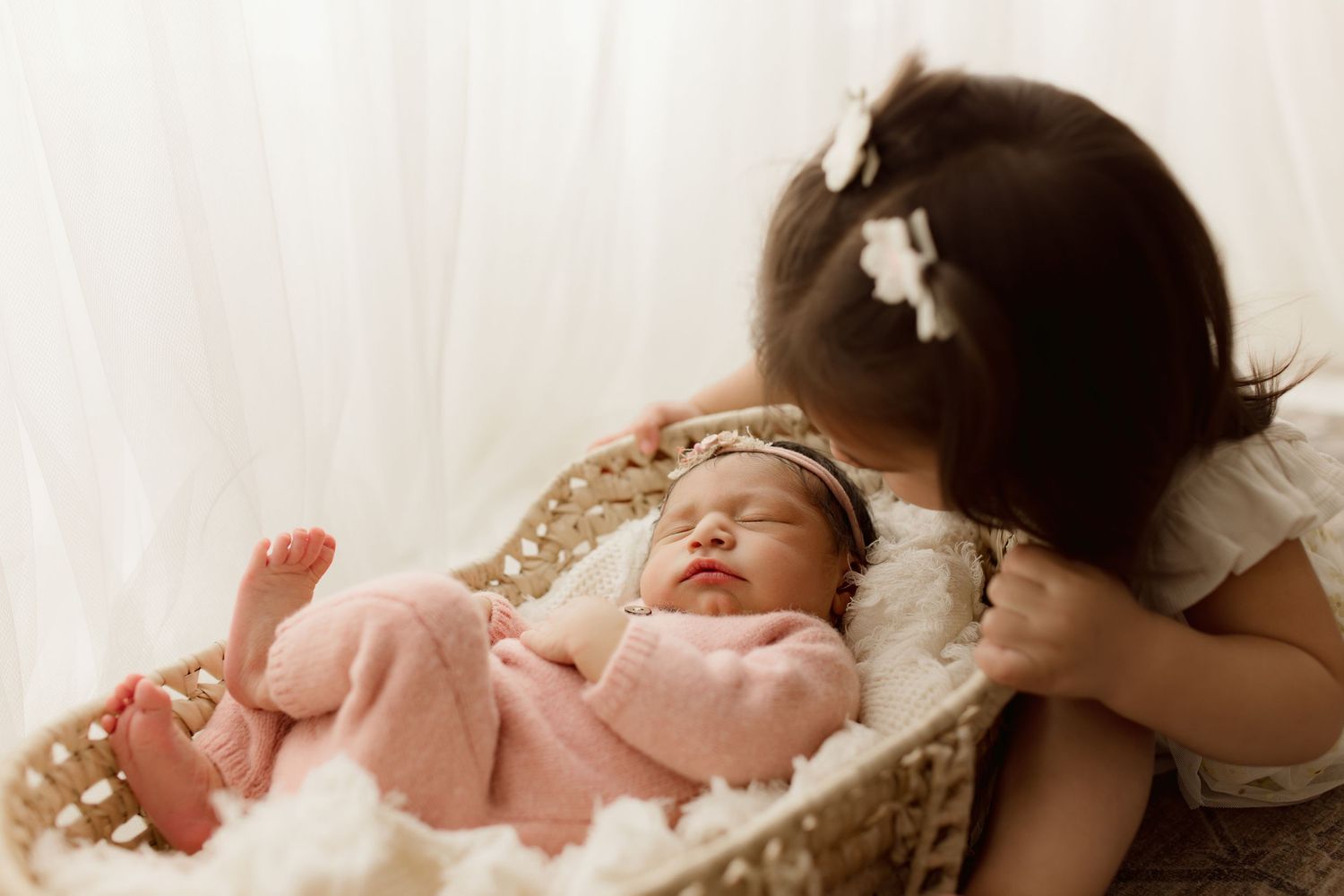 A sleeping newborn baby wearing pink lies in a woven basket while being watched by someone beside them.