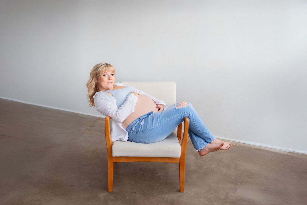 A pregnant woman in blue jeans and light top poses lying on a wooden chair with legs over chair. Studio maternity photoshoot