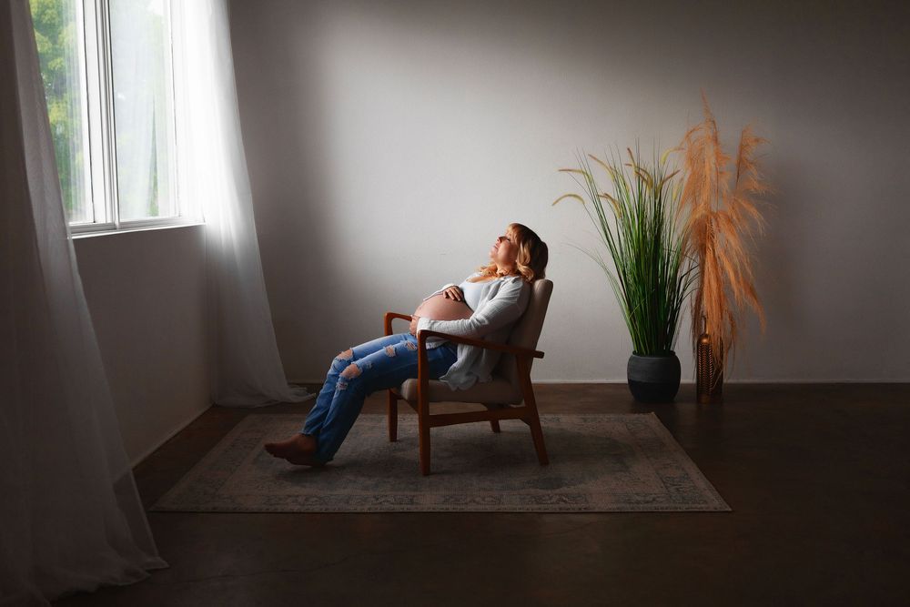 A pregnant woman relaxes in a brown armchair beside a tall decorative grass plant in a minimalist room with moody natural light.