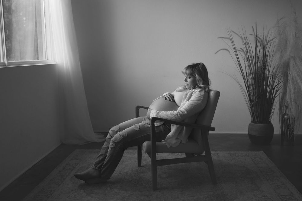 A pregnant woman reclines in a cozy corner armchair beside a window with natural lighting and a potted plant. Black and white photo.