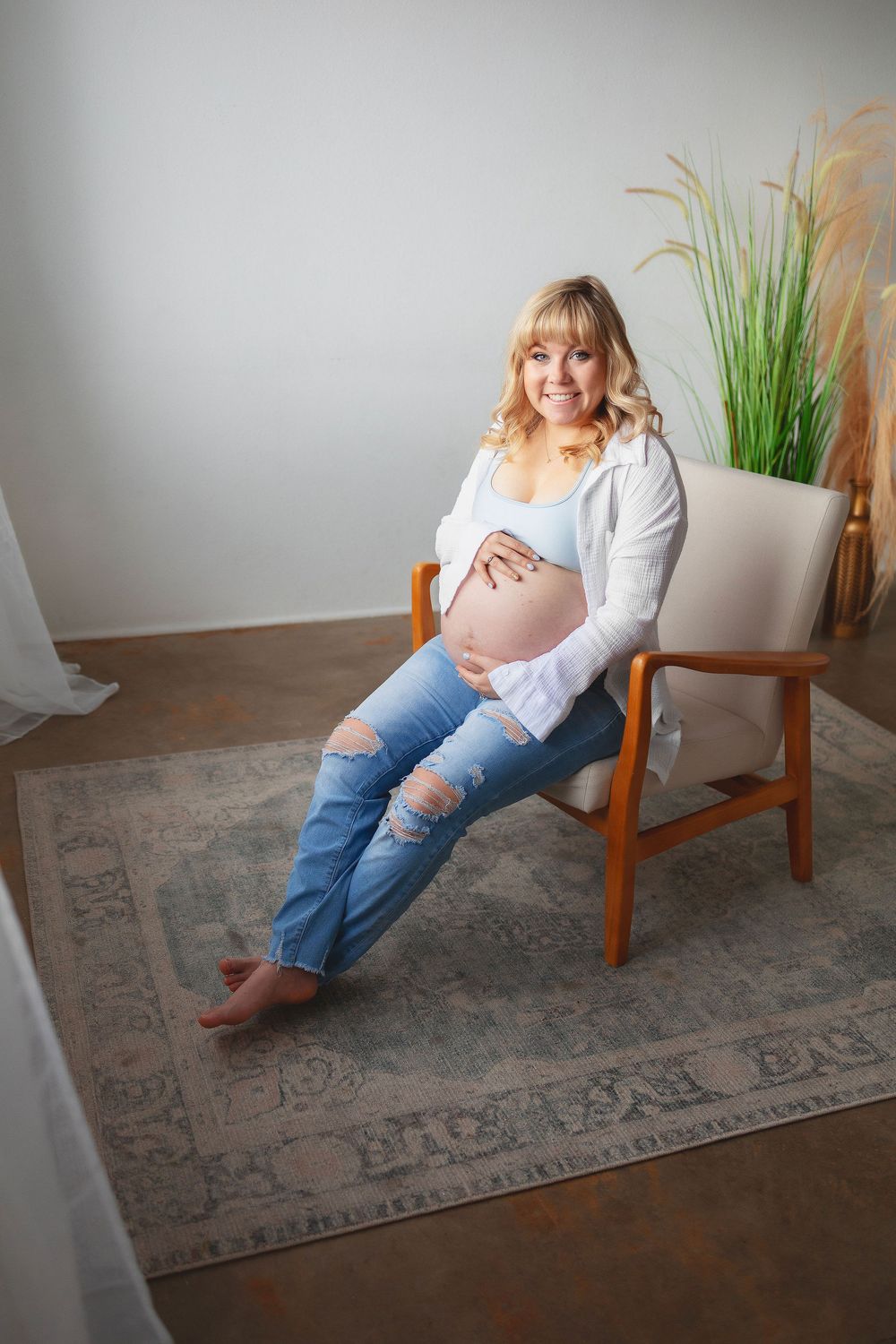 A pregnant woman relaxes in a wooden chair on a patterned area rug, with decorative plants in the background.