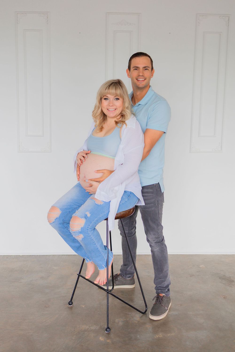 Expecting couple poses together for maternity photo, with the woman sitting on a stool wearing ripped jeans and a gray cardigan.