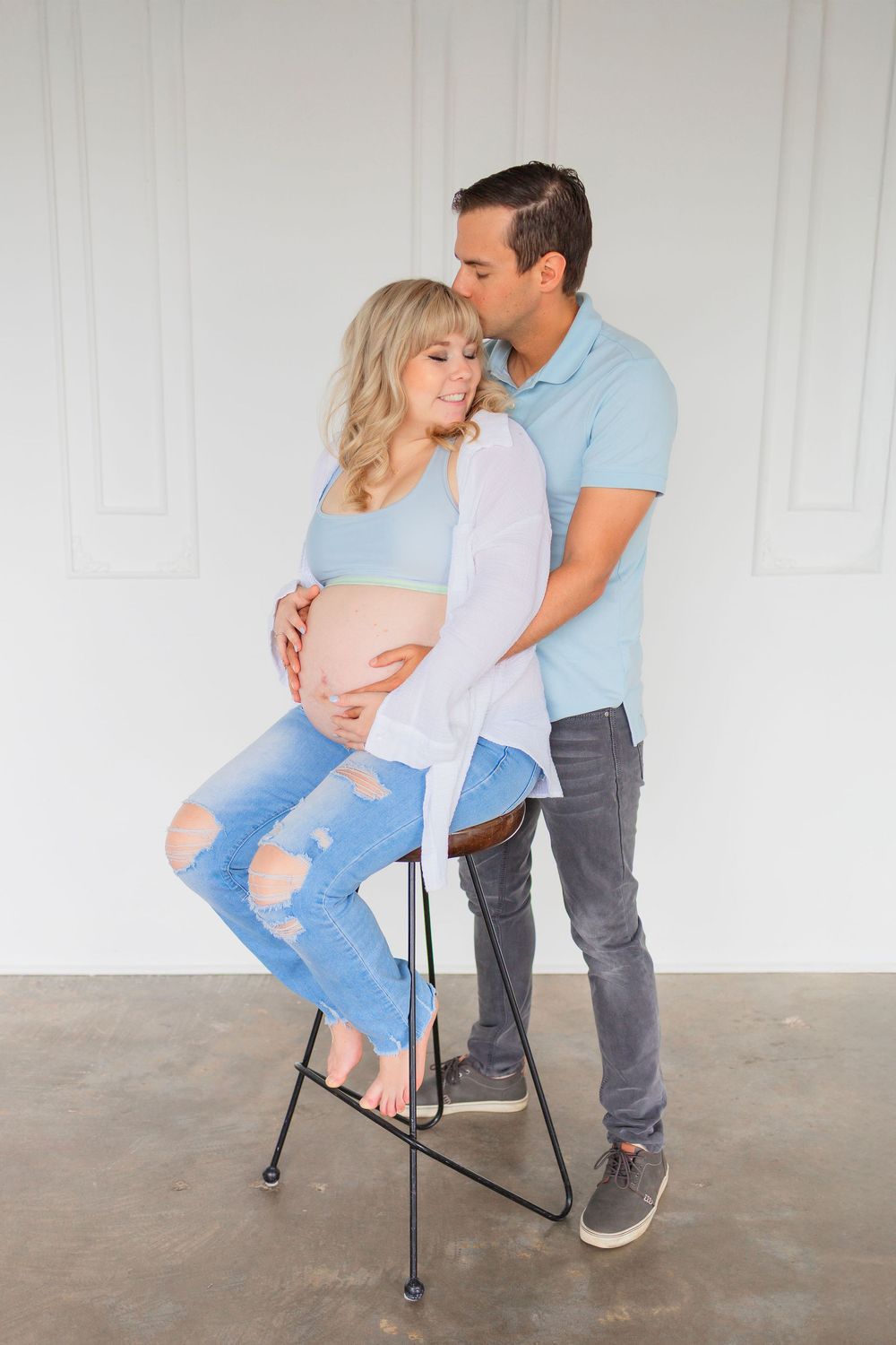 Expecting couple seated against white wall wearing coordinating blue and white outfits during maternity photoshoot.