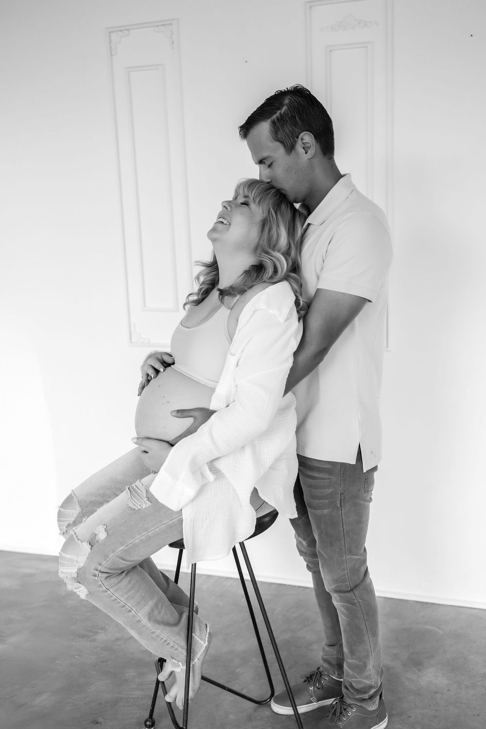 Black and white photo of two people embracing in a tender moment while one sits on a chair against a minimalist white wall.