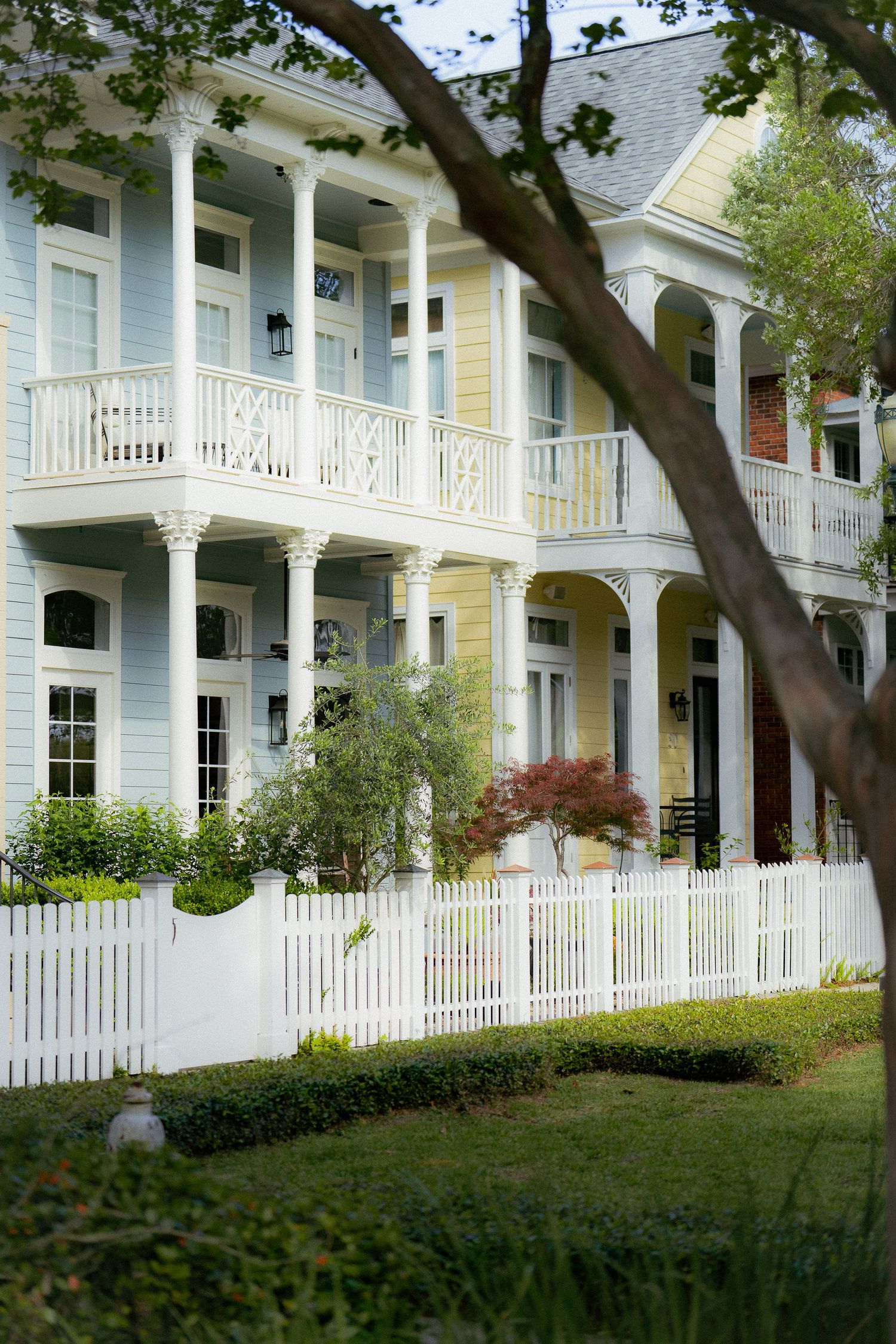 A charming Southern style house with white columns, double-story porches, and a white picket fence in front of a manicured lawn.
